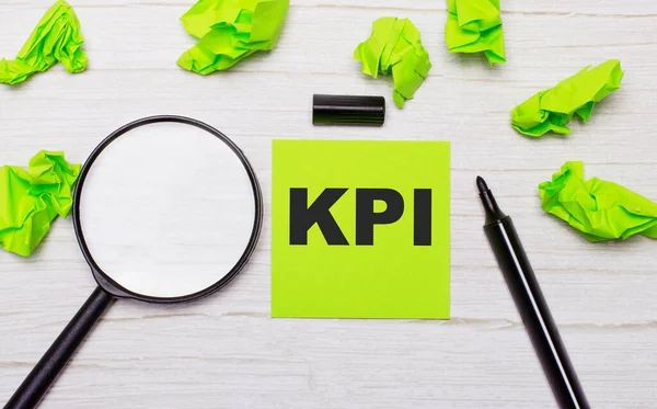 Слово KPI написано на зеленой липкой ноте рядом с лупой и черным маркером на деревянном столе — стоковое фото