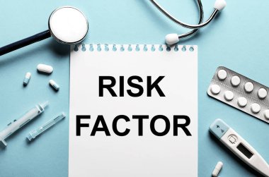 RISK FACTOR kelimesi beyaz bir not defterine steteskop, şırınga, elektronik termometre ve hapların yanında mavi bir arkaplan üzerine yazılmıştır. Tıbbi konsept