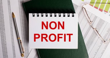 Masaüstünde raporlar, gözlükler, bir kalem, yeşil bir günlük ve içinde NON PROFIT yazan beyaz bir defter var. İş yeri, yakın çekim. İş konsepti