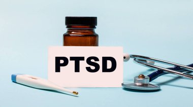 Mavi arka planda haplar için bir şişe steteskop, elektronik termometre ve üzerinde PTSD yazan bir kart var. Tıbbi konsept