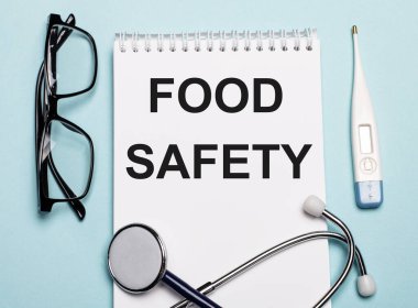 Stetoskop, gözlük ve açık mavi arka planda elektronik termometrenin yanında beyaz bir not defterine yazılan yiyecek güvenliği. Tıbbi konsept.