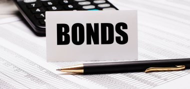 Masanın üzerinde raporlar, bir kalem, bir hesap makinesi ve BONDS yazılı bir beyaz kart var. Odaklanmayın