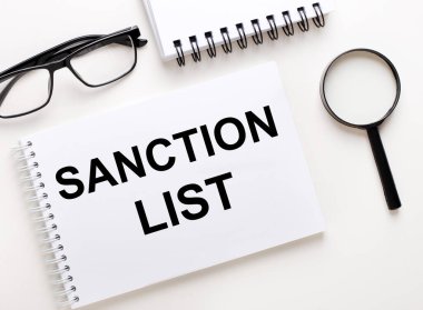 SANCTION LST beyaz bir deftere yazılmıştır defter, siyah çerçeveli gözlük ve bir büyüteç.