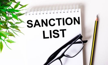 SANCTION LST beyaz bir deftere, siyah çerçeveli bir kalem ve yeşil bir bitkinin yanına yazılmıştır..