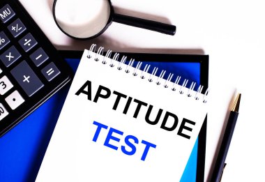 Mavi bir arkaplanda, hesap makinesinin yanında, büyüteç ve kalem, üzerinde APTITUDE TEST yazan bir defter.