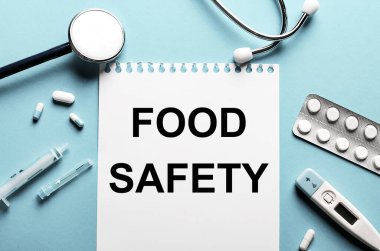 Gıda Güvenliği kelimesi steteskop, şırınga, elektronik termometre ve hapların yanındaki mavi arka planda beyaz bir not defterine yazıldı. Tıbbi konsept