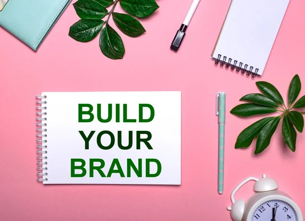 Question BUILD YOUR BRAND is written in green on a white notepad on a pink background surrounded by notepads, pens, white alarm clock and green leaves. Educational concept