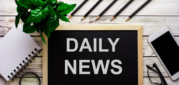 ДЕНЬ ВОПРОСА НОВОСТИ пишется белым цветом на черной доске рядом с телефоном, блокнотом, очками, карандашами и зеленым растением.