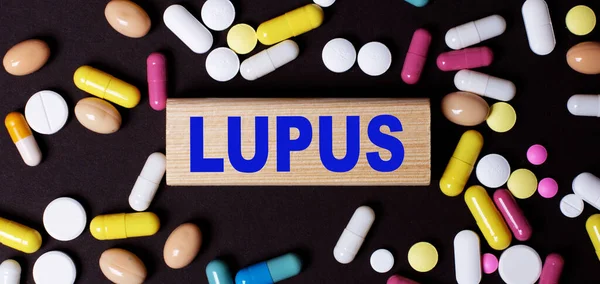 stock image On a dark background, multi-colored pills and the word LUPUS on a wooden block. Medical concept