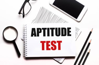 Beyaz bir arka plan gözlüğü, bir büyüteç, kalemler, bir akıllı telefon ve üzerinde APTITUDE TEST yazan bir defter.