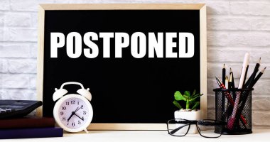 POSTPONED kelimesi beyaz alarm saati, gözlük, saksı bitkisi ve kalem standının yanındaki tahtaya yazılmıştır..