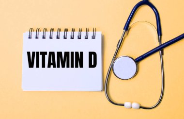 Bej bir arka planda, steteskop ve üzerinde VITAMIN D. yazan beyaz bir not defteri. Tıbbi konsept.