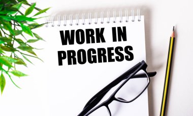 Works in PROGRESS beyaz bir deftere siyah çerçeveli bir kalem ve yeşil bir bitkinin yanında yazılmıştır..