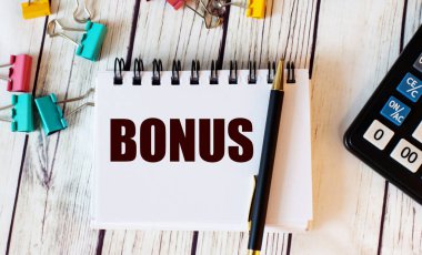 Açık ahşap bir masada bir hesap makinesi, çok renkli ataçlar ve kalemli bir defter ve 