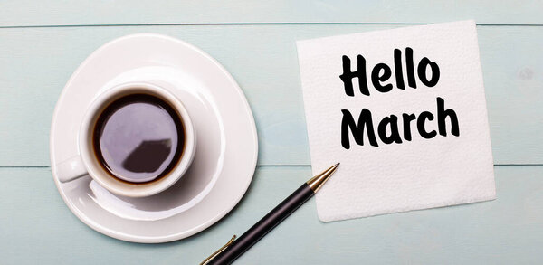 На светло-голубой деревянной дорожке стоит белая чашка кофе, ручка и салфетка с надписью HELLO MARCH.
