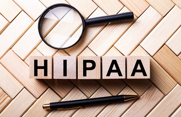 Wooden cubes with the word HIPAA stand on a wooden background between a magnifying glass and a pen