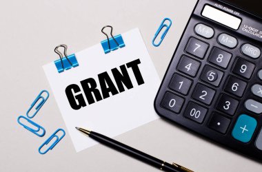 Hafif bir arkaplanda, bir hesap makinesi, bir kalem, mavi ataçlar ve içinde GRANT olan bir kağıt. Yukarıdan görüntüle