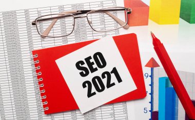 Altın çerçeveli gözlük, kırmızı kalem, renkli masalar ve masaüstünde SEO 2021 yazılı beyaz kart olan kırmızı bir defter. İş konsepti. Yukarıdan görüntüle