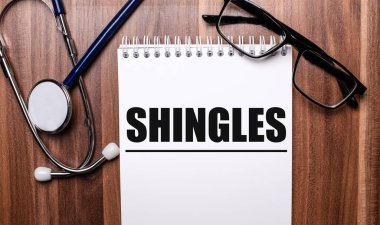 SHINGLES kelimesi, steteskop ve siyah çerçeveli gözlüklerin yanındaki tahta bir arka planda beyaz kağıda yazılmıştır. Tıbbi konsept