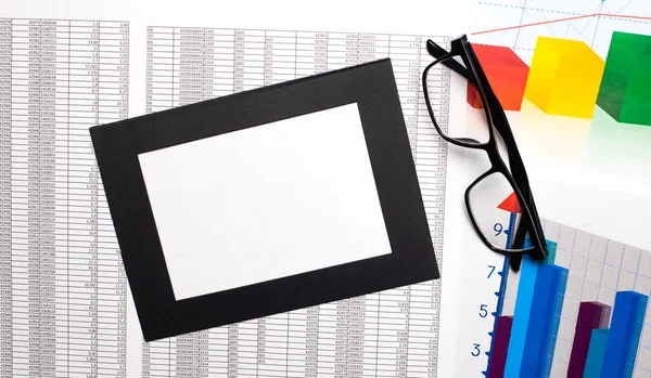 On the desktop are color graphics, black-framed glasses and a blank sheet of white paper in a black frame. Business concept. Top view with copy space