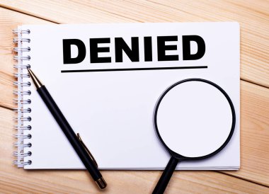 Hafif ahşap bir arka planda bir kalem, bir büyüteç ve üzerinde DENIED yazan bir defter yatar.