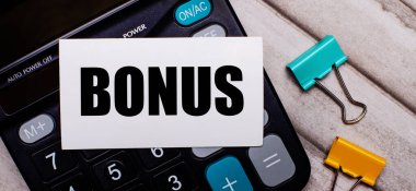 Hesap makinesi, çok renkli ataçlar ve açık tahta bir masa üzerinde BONUS yazan beyaz bir kart. Kırtasiye ve hesap makinesi.