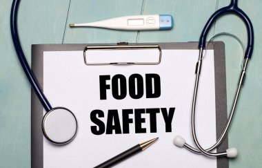 Açık mavi ahşap arka planda Gıda Güvenliği, stetoskop, elektronik termometre ve kalem etiketli bir kağıt var. Tıbbi konsept