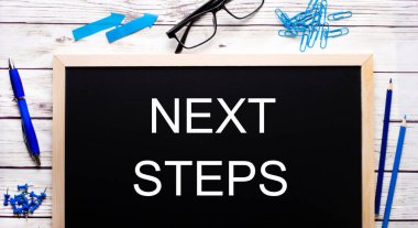 Mavi ataçlar, kalemler ve kalemlerin yanına siyah bir not panosuna yazılmış NEXT STEPS.