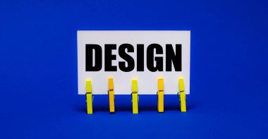 Sarı mandallar üzerinde parlak mavi bir arkaplanda DESIGN yazılı beyaz bir kart var.