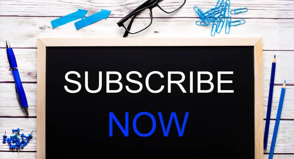 SUBSCRIBE NOW written on a black note-board next to blue paper clips, pencils and a pen.