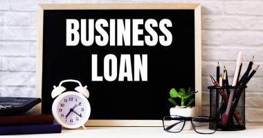 BUSINESS kredisi kelimesi beyaz alarm saati, gözlük, saksı bitkisi ve kalem standının yanındaki tahtada yazılıdır..