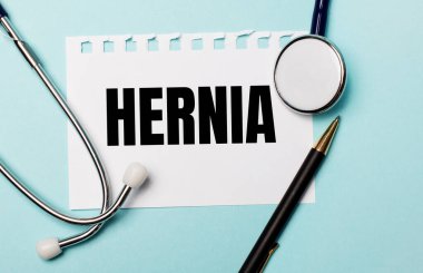 Açık mavi bir arkaplanda, steteskop, bir kalem ve üzerinde HERNIA yazan bir kağıt. Tıbbi konsept