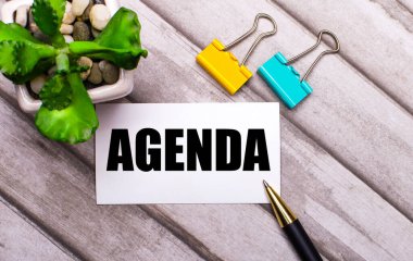 Ahşap bir arka planda, üzerinde AGENDA yazılı beyaz bir kart, sarı ve yeşil ataçlar ve saksıda bir bitki. Yukarıdan görüntüle