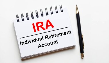 On a light background, a white notebook with are words IRA Individual Retirement Account and a pen.