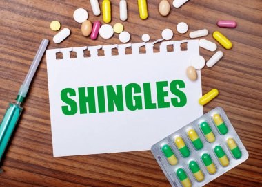 On a wooden table, a syringe, pills and a sheet of paper with the inscription SHINGLES. Medical concept