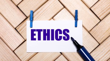 Ahşap bir arka planda mavi mandallar ve mavi işaretli ETHICS yazılı beyaz bir kart var. Yukarıdan görüntüle