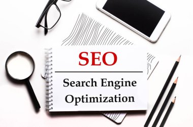 Beyaz bir arka plan gözlüğü, bir büyüteç, kalemler, bir akıllı telefon ve SEO Arama Motoru En İyimser metni olan bir defter üzerinde