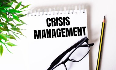 CRISIS MANAGEMENT bir kalem, siyah çerçeveli gözlük ve yeşil bir bitkinin yanında beyaz bir deftere yazılmıştır..