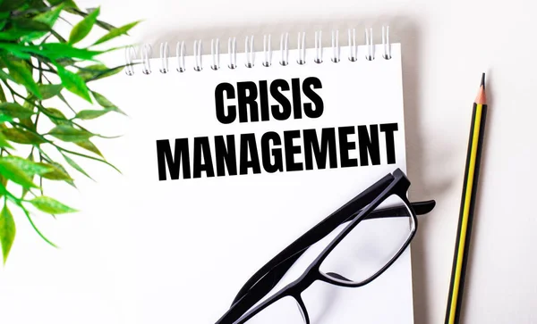 CRISIS MANAGEMENT is written in a white notebook next to a pencil, black-framed glasses and a green plant.