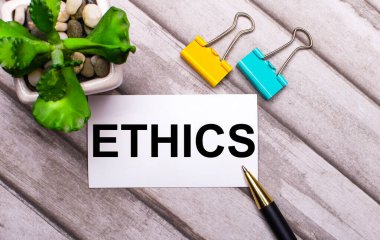Ahşap bir arka planda ETHICS yazılı beyaz bir kart, sarı ve yeşil ataçlar ve saksıda bir bitki. Yukarıdan görüntüle