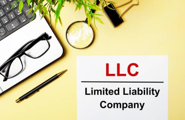 LLC Limited Limited Borbility Company, açık sarı arka planda, dizüstü bilgisayar, kalem, büyüteç, gözlük ve yeşil bitkinin yanında beyaz bir kağıt parçasına kırmızı ile yazılmıştır..