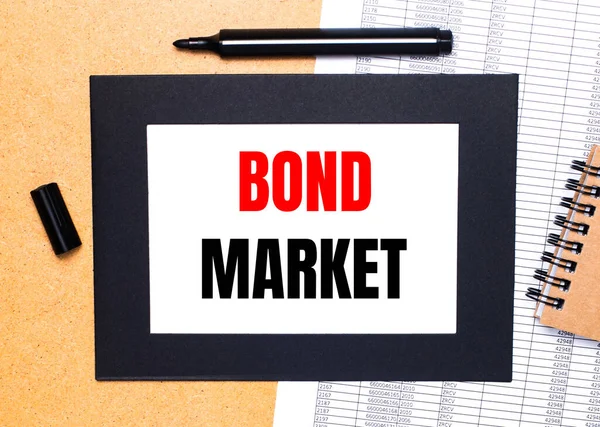 On a wooden table, there is a black open marker, a brown notepad and a sheet of paper in a black frame with the text BOND MARKET. View from above.