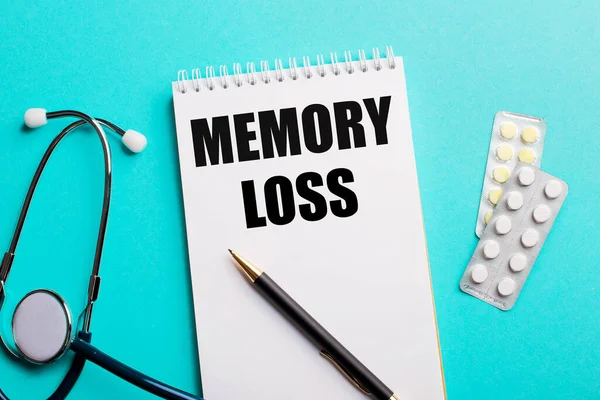MEMORY LOSS written in a white notepad near a stethoscope, pens and pills on a light blue background. Medical concept