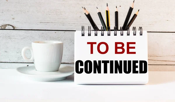 The words TO BE CONTINUED is written in a white notepad near a white cup of coffee on a light background