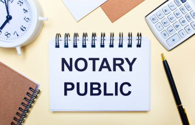 Hafif bir arkaplanda, bir beyaz alarm saati, bir hesap makinesi, bir kalem ve üzerinde NOTARY PUBLIC yazan bir defter. Düz yatıyordu
