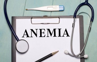 Açık mavi ahşap arka planda ANEMIA etiketli bir kağıt, stetoskop, bir elektronik termometre ve bir kalem var. Tıbbi konsept