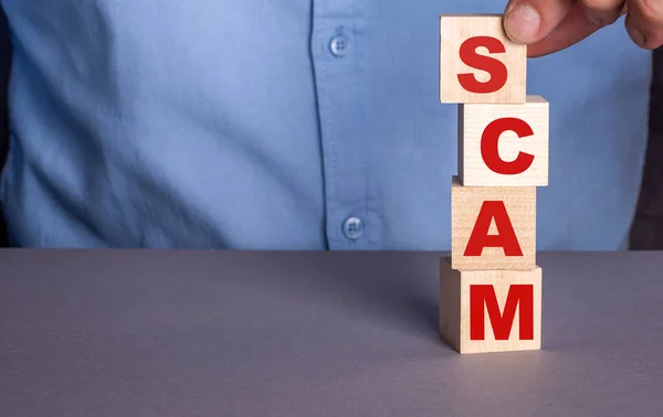 Mavi gömlekli bir adam dikey olarak tahta küplerden SCAM sözcüğü yazıyor.