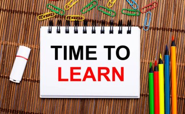 Ahşap bir masada, çok renkli kalemler, ataçlar, beyaz bir flaş bellek ve Time to LeARN metninin olduğu bir defter. Düz yatıyordu