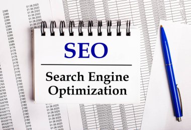 Masanın üzerinde, üzerinde SEO Arama Motoru İyimserleştirmesi yazan mavi bir kalem ve bir defter bulunan tablolar ve raporlar var.