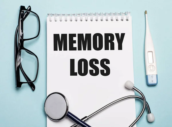 MEMORY LOSS written on a white notepad next to a stethoscope, goggles, and an electronic thermometer on a light blue background. Medical concept.